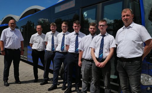 Acht Männer bekleidet mit weißen Hemden und dunklen Hosen stehen seitlich vor einem Bus