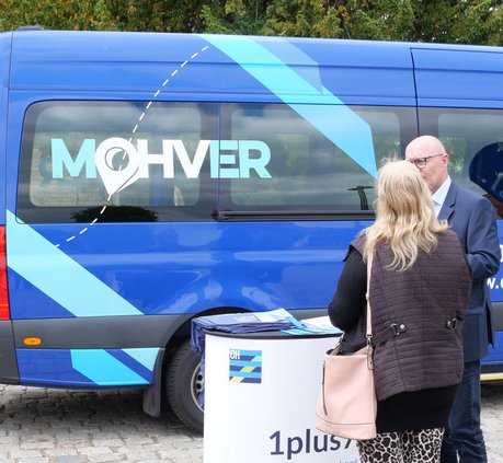 Seitenansicht des MOHVER, davor steht ein Infocounter, an dem sich zwei Leute unterhalten.
