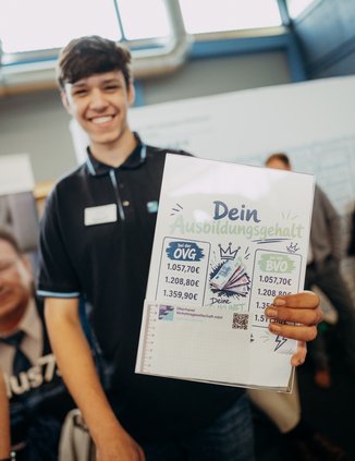 Ein junger Mann hält eine Übersicht mit den Ausbildungsgehältern ins Bild.